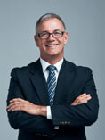 Smiling, professional-looking grey haired man with glasses in dark suit with arms crossed.