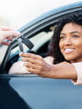 Black woman in driver's seat of black car and someone is handing her keys through the driver's window.