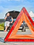 Man leaning on rear bumper of car with trunk lid open in background; orange caution triangle in foreground.