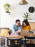 Couple at kitchen table talking in front of laptop screen.