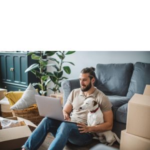 Man in apartment on laptop next to his dog and boxes everywhere.